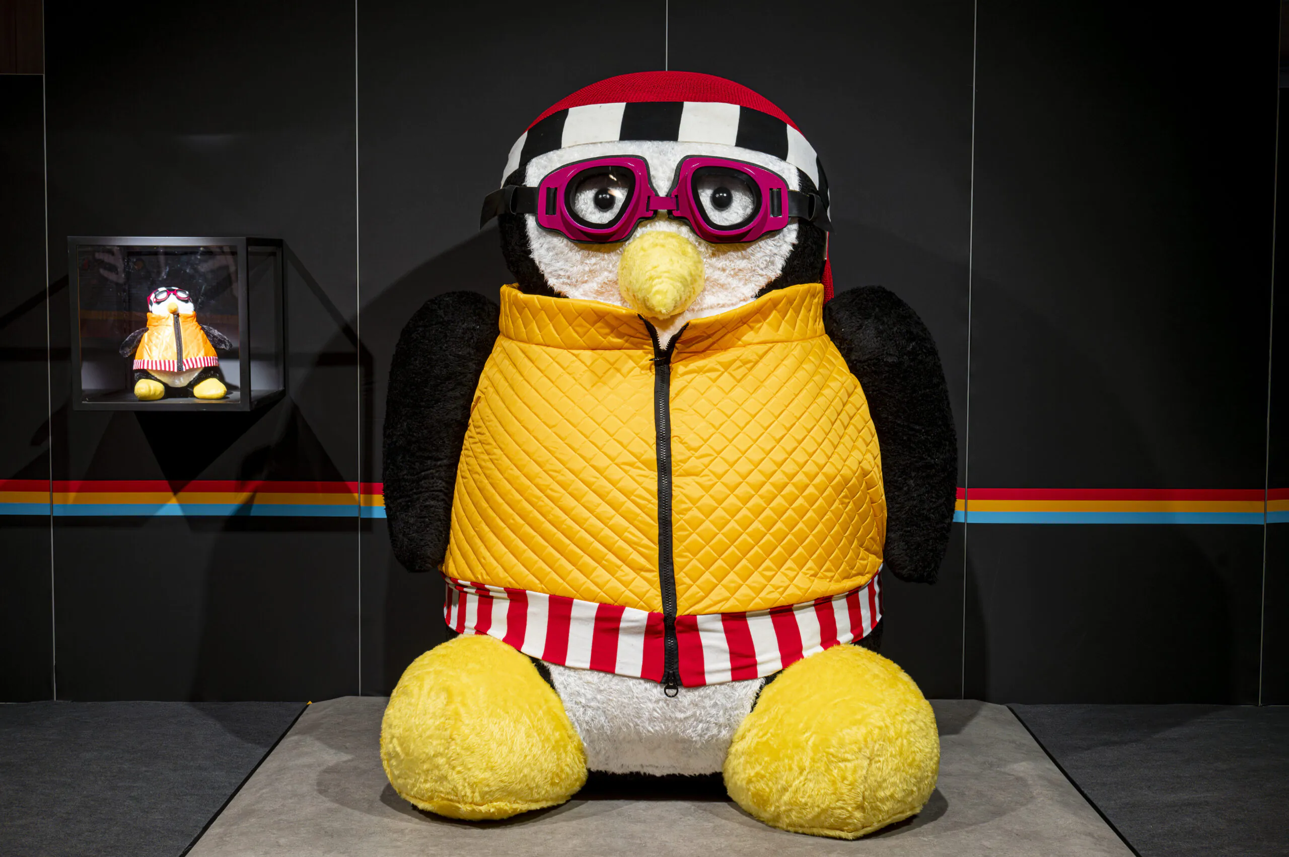 La peluche Hugsy représente un pingouin avec lunettes, écharpe et veste jaune.