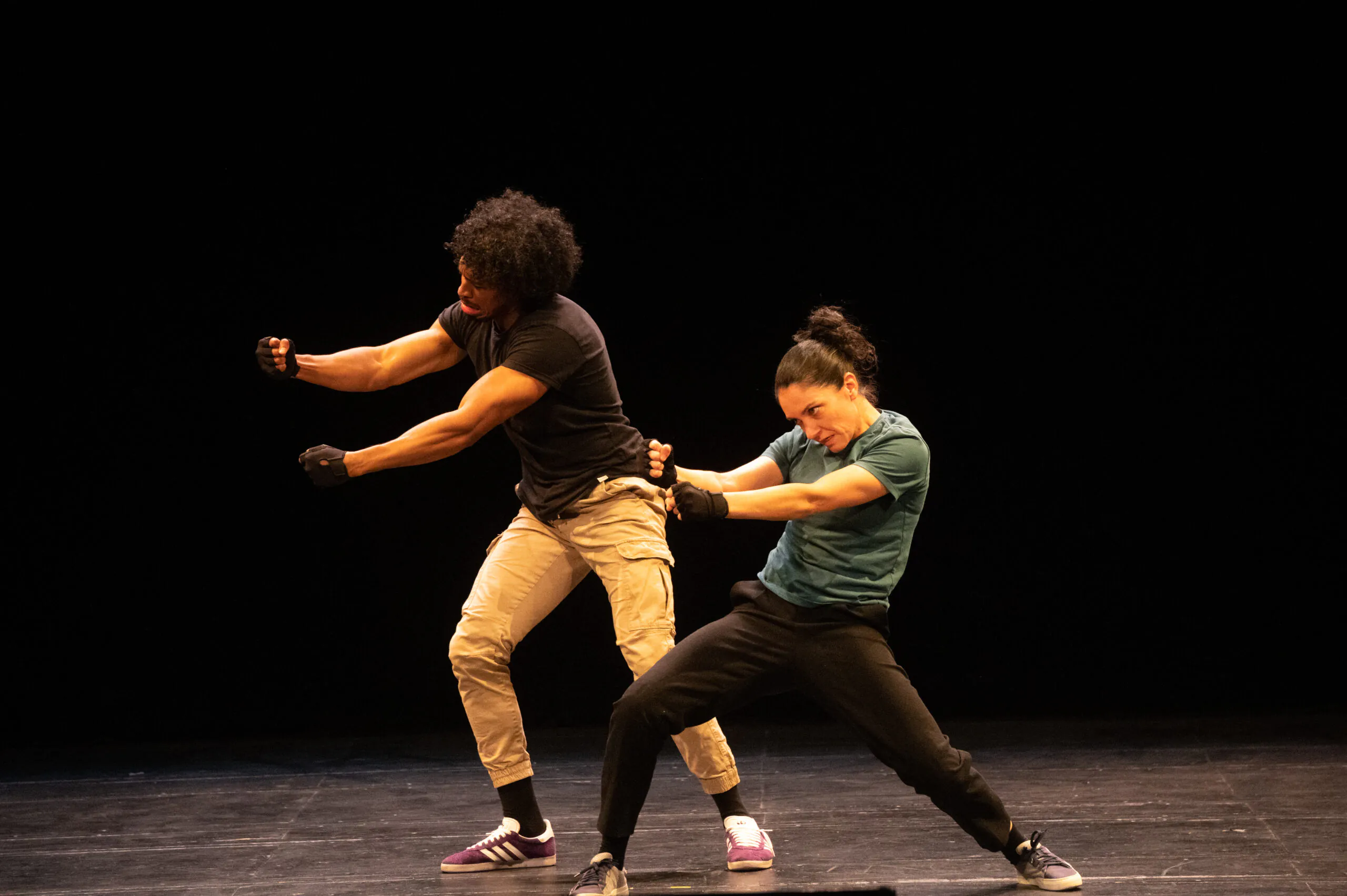 Sur fond noir, un danseur et une danseuse en baskets, t-shirts et pantalon, les bras tendus côté jardin, genoux fléchis et tête regardant leurs mains.