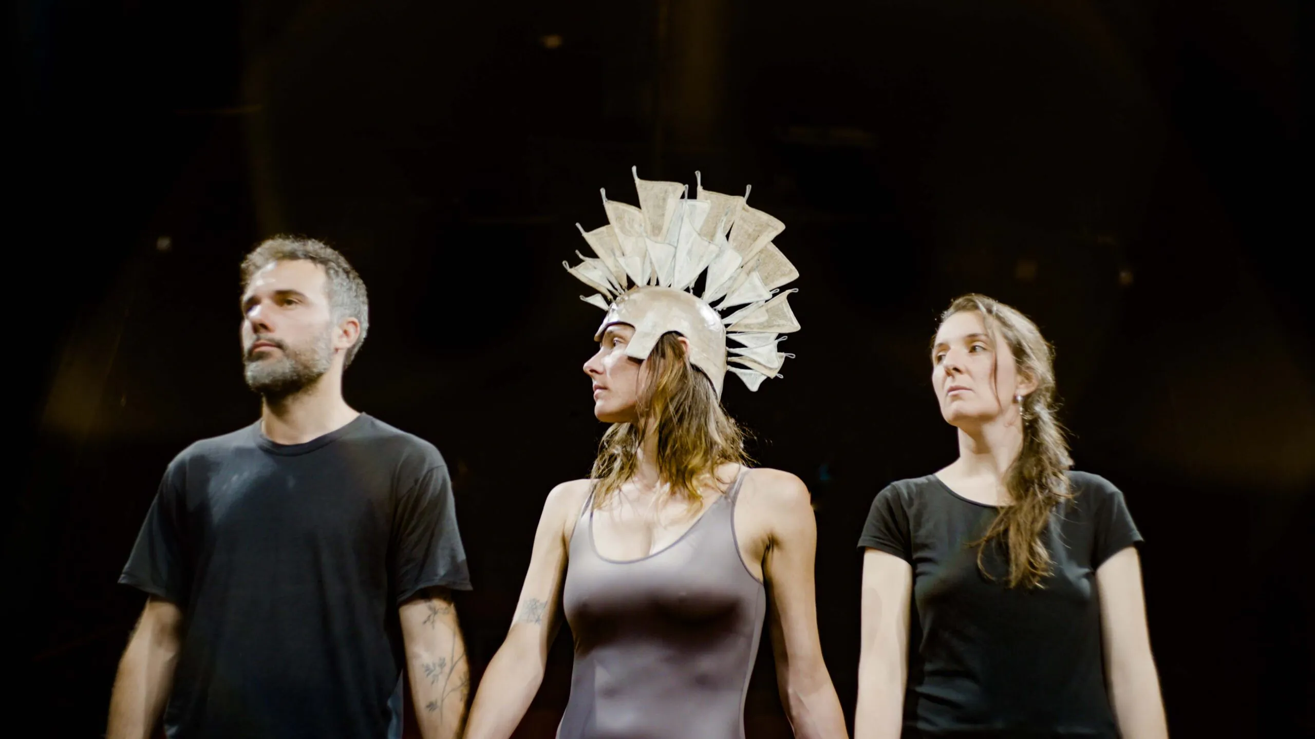 Image sur fond noir avec un danseur et deux danseuses qui se tiennent par la main. La danseuse du milieu a une couronne façon casque de guerrière.